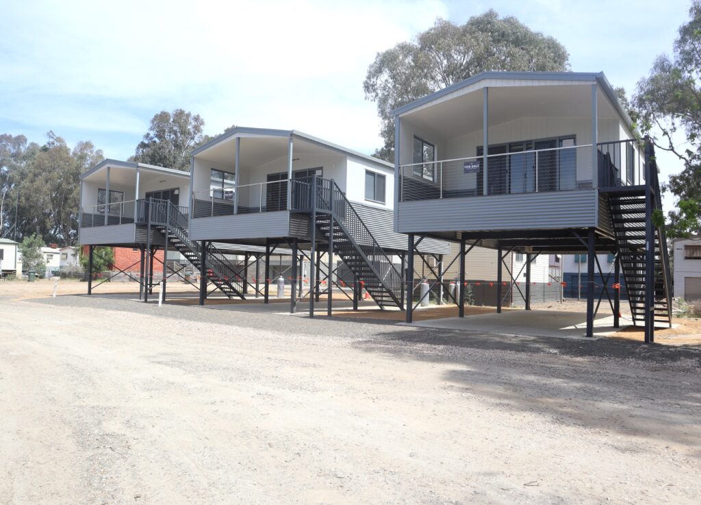 Three elevated modern houses on stilts with stairs leading to the entrances. The structures have a sleek, metallic exterior and are situated on a gravel lot, surrounded by tall trees in the background.