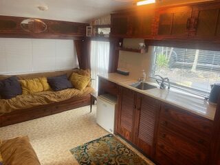 Interior of a cozy trailer with wood-paneled walls, featuring a yellow sofa with gray pillows on the left, a kitchenette with sink and cabinets on the right, and bright windows letting in natural light. A small rug is on the light-toned floor.