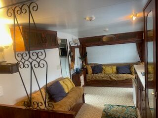 Interior of a cozy RV living space with two brown sofas, each adorned with blue and beige cushions. The room features wood paneling, a small lamp, a patterned carpet, and a decorative metal partition. Natural light filters in through a window.