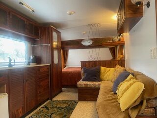 Cozy interior of a wooden RV featuring a beige sofa with yellow and blue cushions, a kitchenette with cabinets, and a bed in the back. A window with blinds provides natural light, and a small carpet adds to the decor.