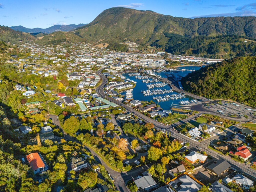 Aerial view of Picton, a coastal town framed by lush green hills, with a marina dotted with boats, winding roads, and scattered buildings. The landscape includes residential areas and open spaces under a blue sky adorned with clouds.