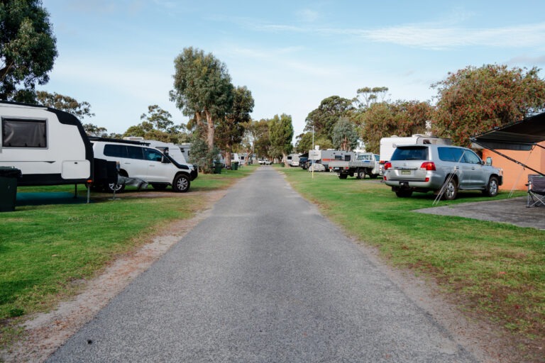 A paved road winds through the campsite, flanked by parked RVs, caravans, and vehicles on grassy areas. Trees line the path under a clear sky. Tables and chairs are set up near some RVs and caravans, inviting you to enjoy the camping experience.