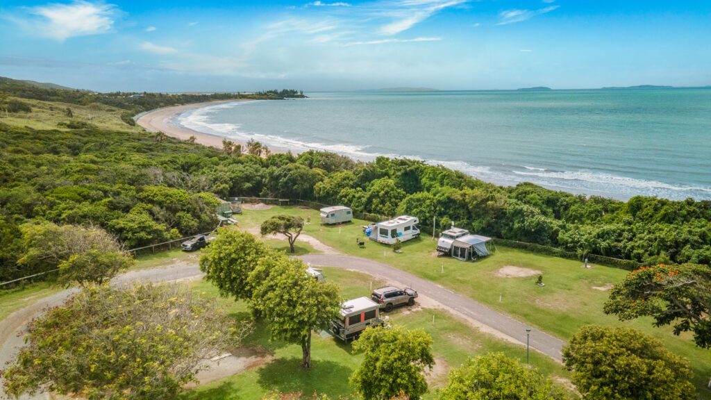 Aerial view of a coastal camping site in Yeppoon, adjacent to a sandy beach and ocean. Several RVs and campers are parked on a grassy area surrounded by trees and shrubs. The shoreline curves along the left, with gentle waves and a clear blue sky overhead.