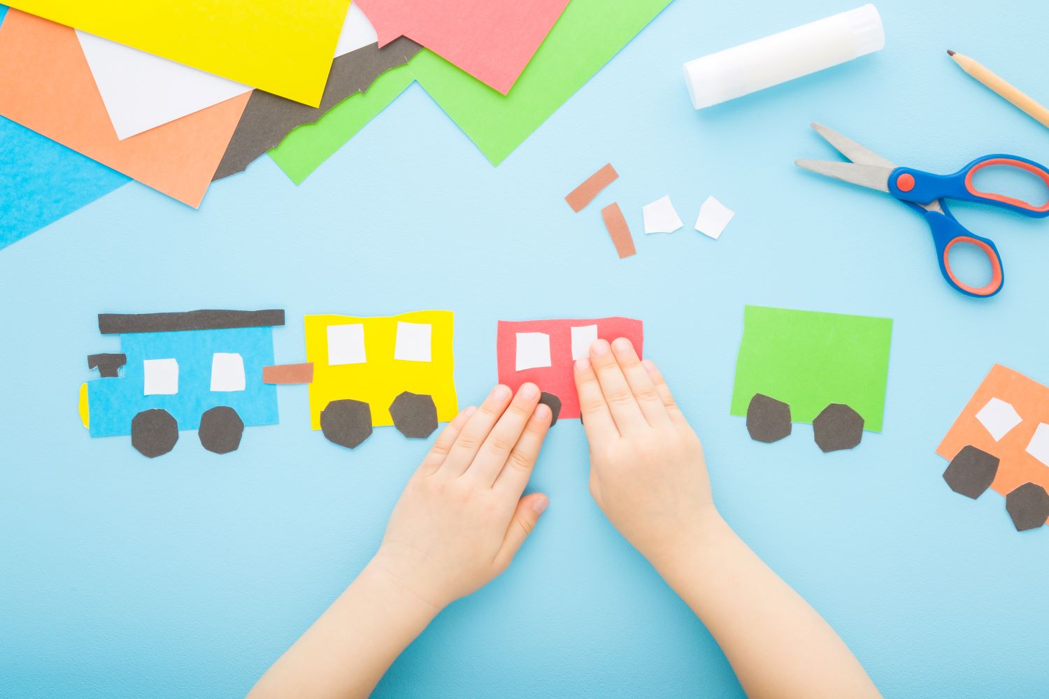 Child's hands assembling a colorful paper train on a blue background, surrounded by sheets of colored paper, scissors, a pencil, and glue. The train consists of a blue, yellow, red, and green car.