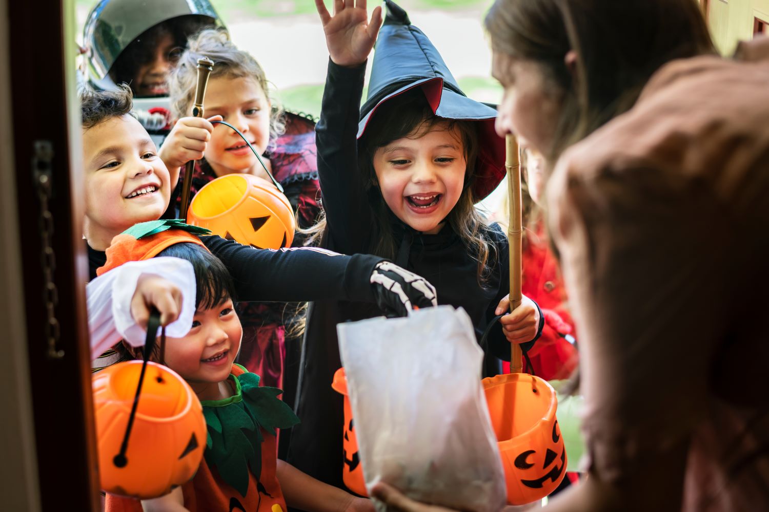 A group of excited children dressed in various Halloween costumes hold out pumpkin-shaped buckets for candy from a person at a door. Smiling and joyful, the kids eagerly await their treats during trick-or-treating.