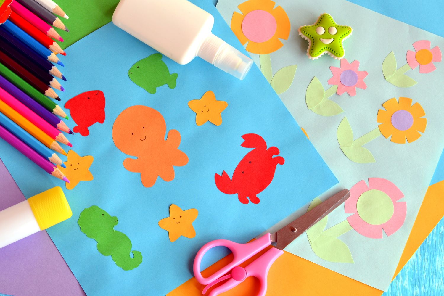 Colorful crafting materials on a table include cut-out sea creature shapes, a star-shaped eraser, colored pencils, glue, and scissors. There are also sheets of paper with flower and leaf designs.