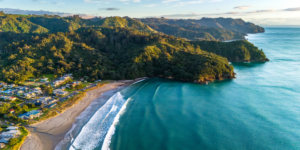 Aerial view of a coastal landscape featuring a sandy beach curving along the shoreline, surrounded by lush green hills. The perfect place to unwind with waves gently wash onto the shore, while houses are nestled among the trees near the beach. The ocean stretches to the horizon.