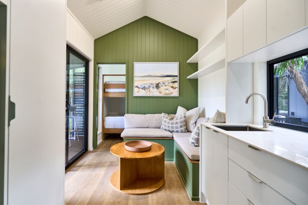 A modern interior inspired by Yallingup Beach showcases a small living space with green paneled walls, a cozy sectional sofa, and a round wooden coffee table. The room features sliding glass doors, a minimalist kitchen area, and a bedroom visible through an open door.