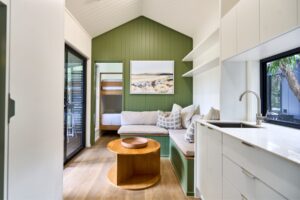 A modern interior inspired by Yallingup Beach showcases a small living space with green paneled walls, a cozy sectional sofa, and a round wooden coffee table. The room features sliding glass doors, a minimalist kitchen area, and a bedroom visible through an open door.