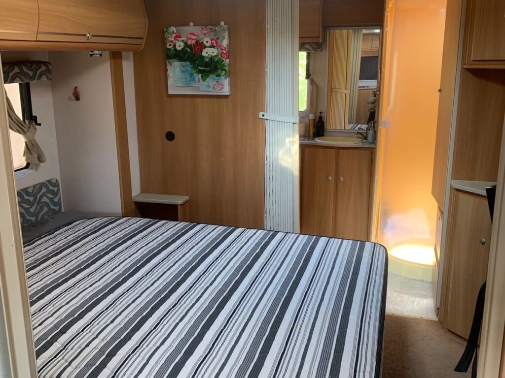 Interior of a camper van featuring a striped bed, wooden cabinets, a floral painting on the wall, and a view of a small bathroom with a shower. Natural light comes in through a skylight.