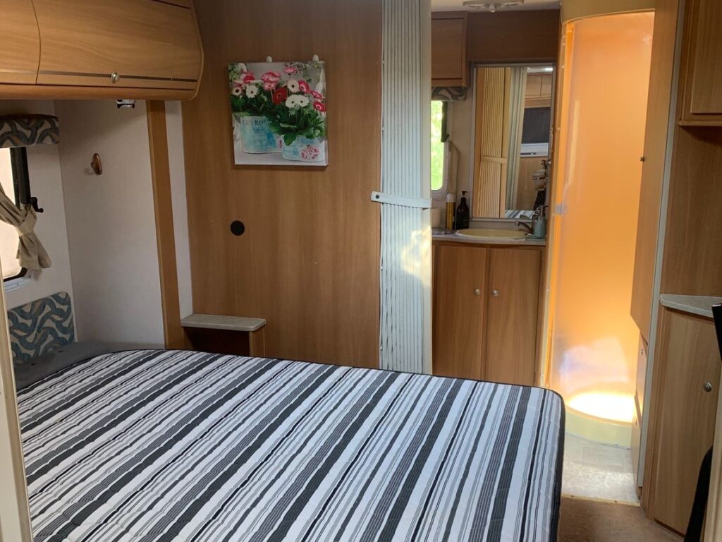 Interior of a camper van featuring a striped bed, wooden cabinets, a floral painting on the wall, and a view of a small bathroom with a shower. Natural light comes in through a skylight.