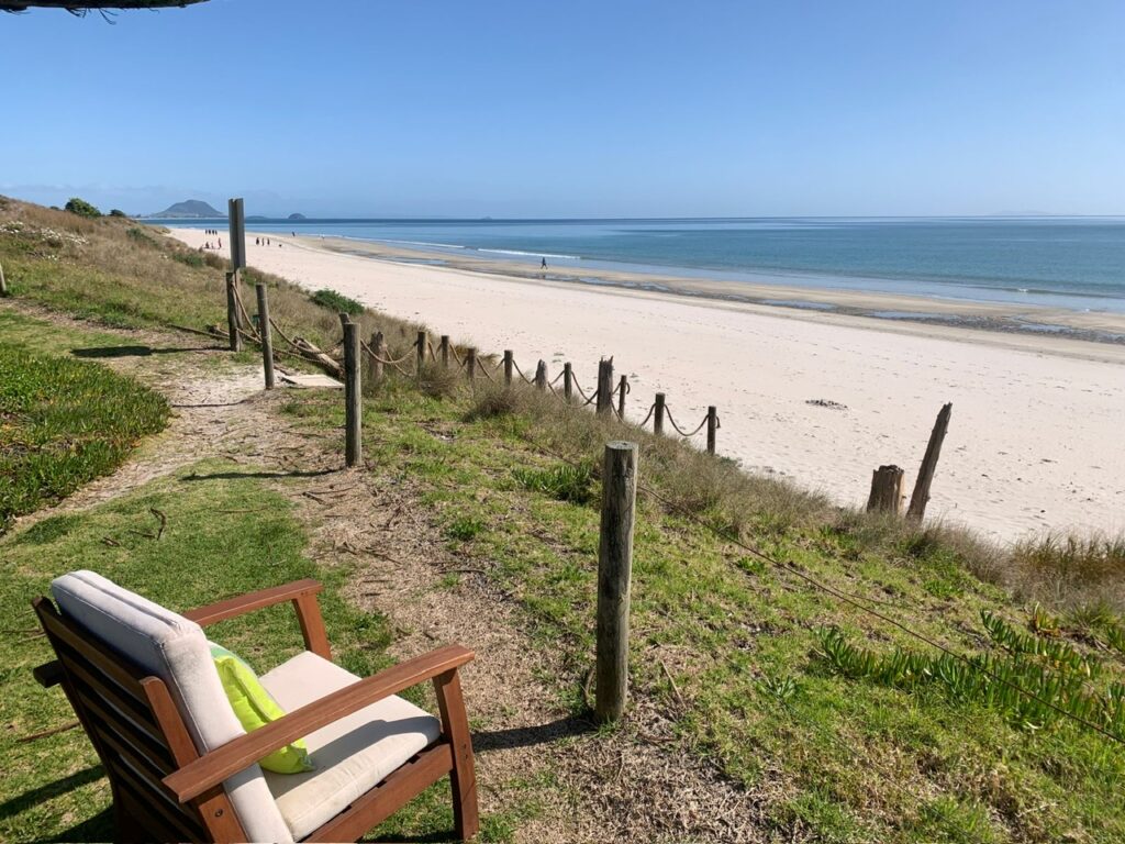 A wooden chair with a cushion sits on a grassy hill overlooking a sandy beach. A few people are walking along the shoreline under a clear blue sky. Wooden posts and rope form a simple barrier along the edge of the hill.