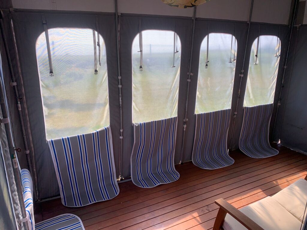 A sunlit room with four mesh windows and blue-striped blinds. The floor features wooden slats, and a wooden chair is partially visible. Light casts patterns through the blinds onto the floor.