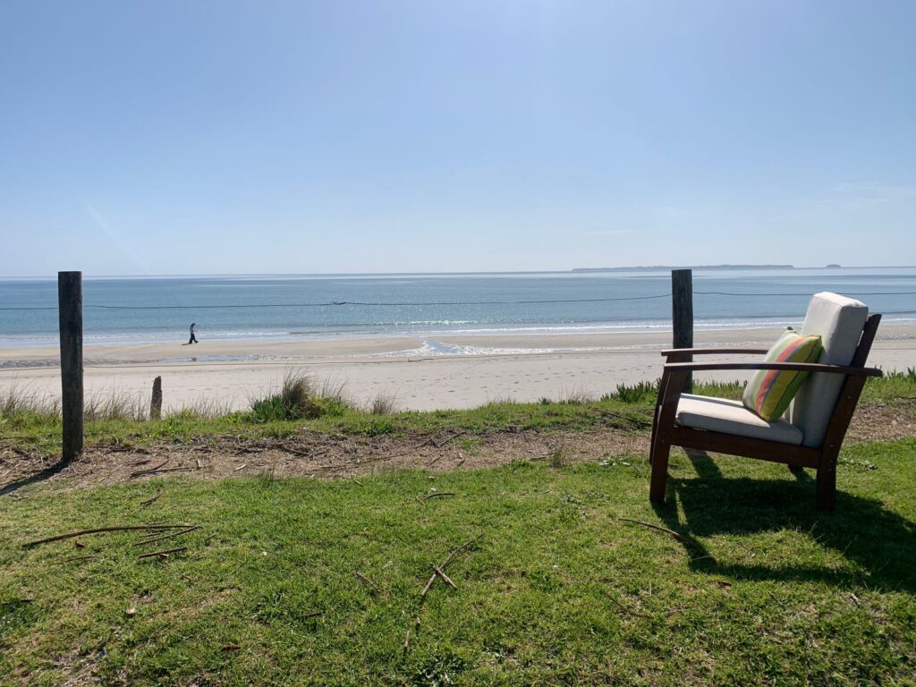 A wooden chair with cushions sits on a grassy area overlooking a sandy beach and calm ocean. A person walks along the shoreline under a clear blue sky. Fence posts line the edge of the grass.