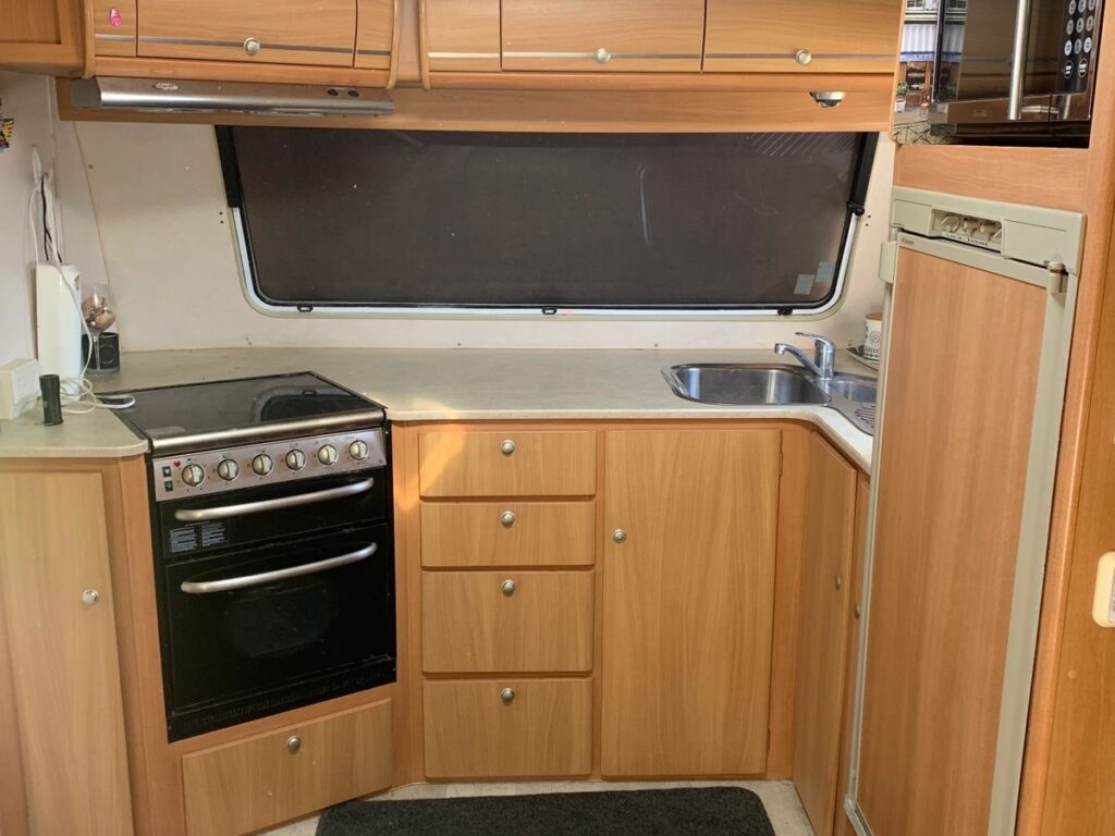 Interior of a camper van kitchen with wooden cabinets, an oven, stove, microwave, sink, and refrigerator. A black mat covers part of the floor, and the space is illuminated by natural light from a window.