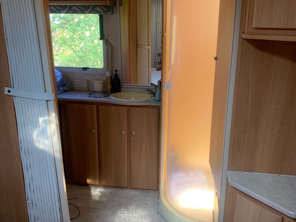 Interior of a caravan with wooden cabinets. On the left, a small kitchenette with a sink is visible beneath a window. On the right, there's a curved, frosted-glass shower. A bed is partially visible in the foreground.