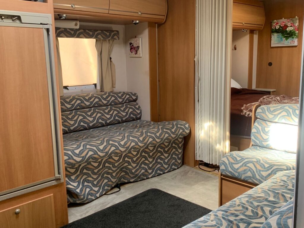 Interior of a camper with wood paneling, featuring a small kitchen area with a microwave, seating with patterned cushions, and a bed in the background. Cozy space with natural light coming through a window.