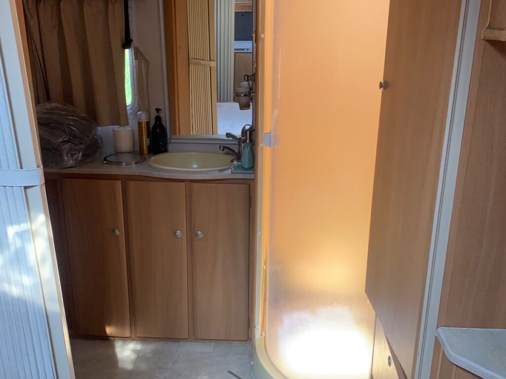 Interior of a compact RV with a wooden finish. The image shows a small sink and counter, an orange-tinted round shower cubicle, and various storage cabinets. Natural light filters through a window, creating a warm ambiance.