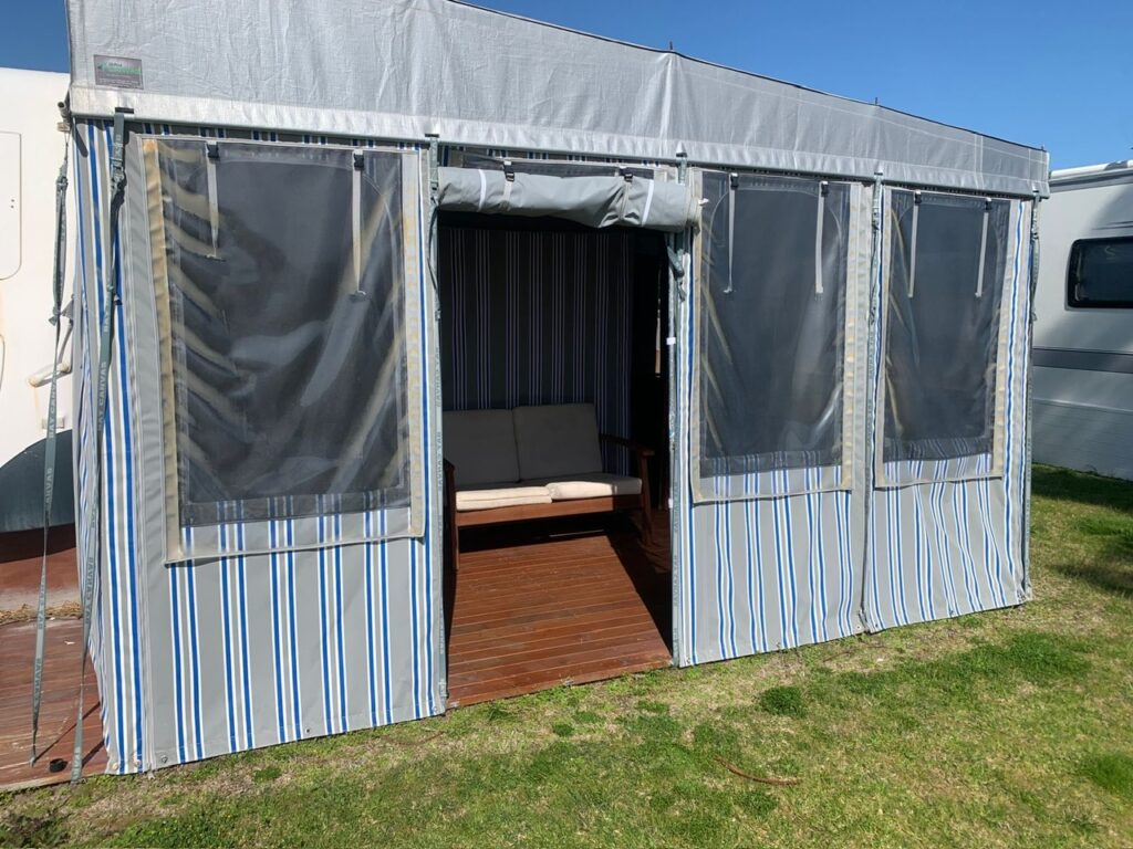 A small outdoor gazebo with blue and white striped fabric walls, featuring a wooden floor and a cushioned bench inside. The structure is set on grass, with a clear blue sky in the background.