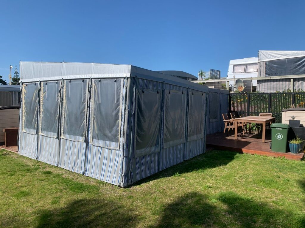 A large tent with transparent panels stands on a grassy lawn under a clear blue sky. Adjacent to the tent, there's a wooden deck with a picnic table and benches. A black fence and buildings are visible in the background.