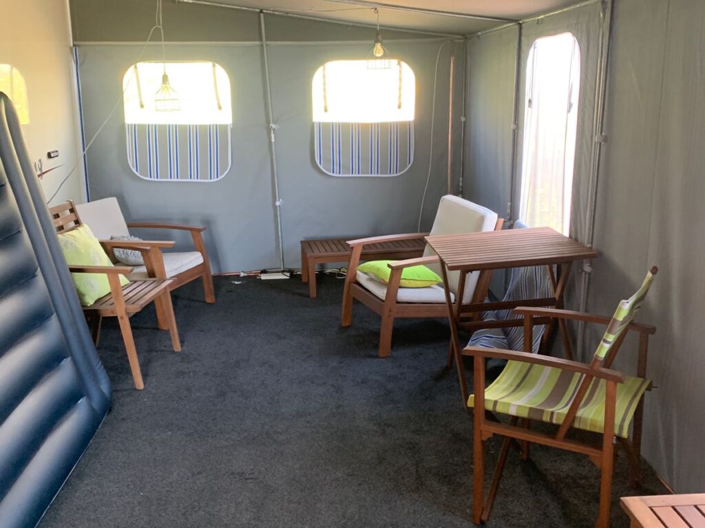 Interior of a tent with dark carpet, featuring wooden chairs with green cushions, a table, and striped fabric panels on the windows. An inflatable mattress leans against the left wall.