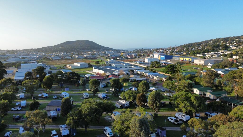 Aerial view of a campground with numerous vehicles, caravans, and tents, surrounded by trees. In the background, there are buildings and a hilly landscape under a clear blue sky.