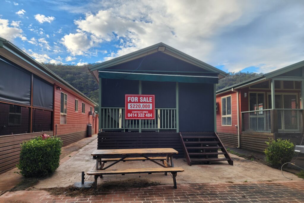 A small blue house is for sale for $220,000, with contact number displayed on a sign. It is situated between two other houses, with a picnic table in front. The sky is partly cloudy, and there are hills in the background.