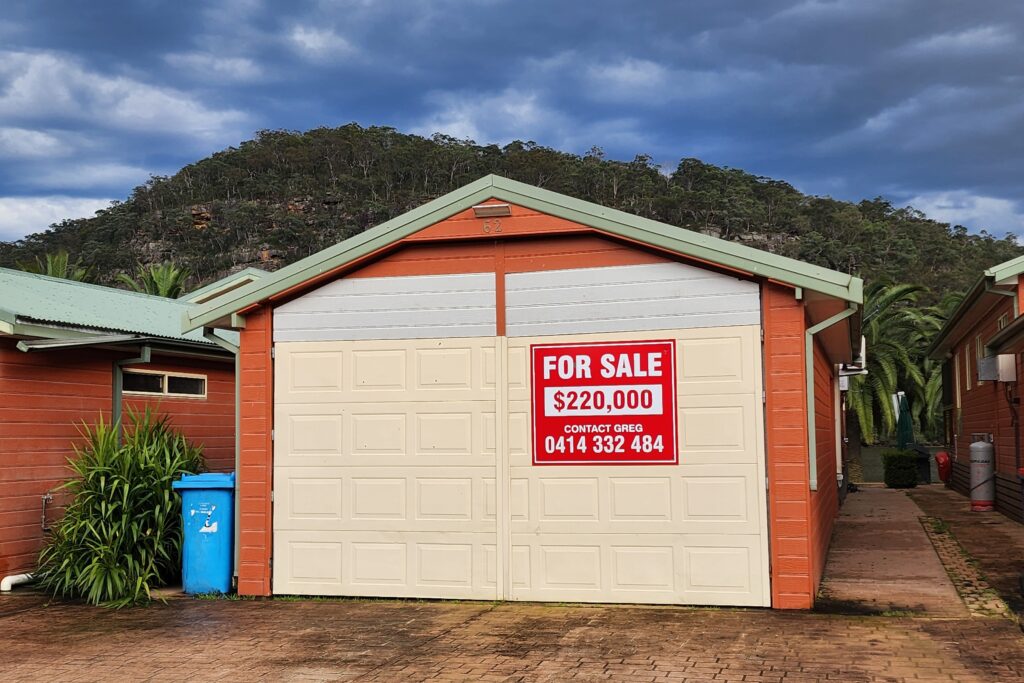 A brown house with a green roof has a large "For Sale" sign on its garage door, listing the price as $220,000 with a contact number. The home is set against a backdrop of lush green hills under a cloudy sky.