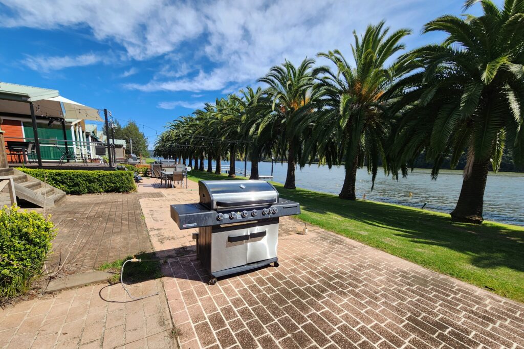 A stainless steel grill is on a brick patio next to a river, surrounded by palm trees and greenery under a blue sky. In the background, there are outdoor seating areas and buildings.