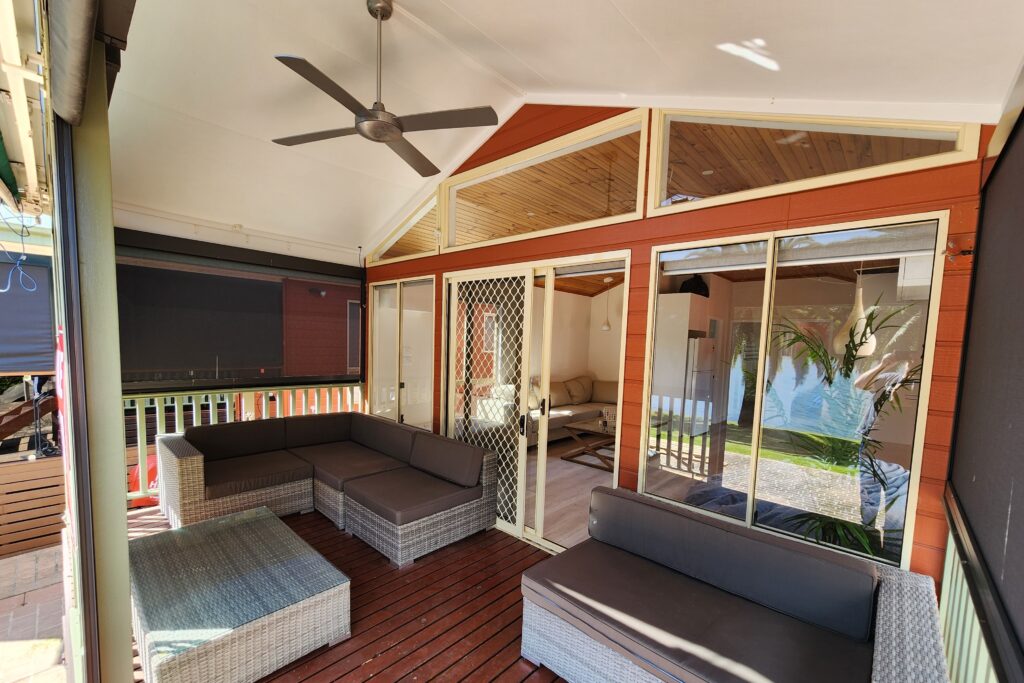 A cozy outdoor patio with wicker furniture and a coffee table on a wooden deck. The space is enclosed with sliding glass doors and features a ceiling fan. Sunlight filters in through the windows, illuminating the light-colored walls and plants outside.
