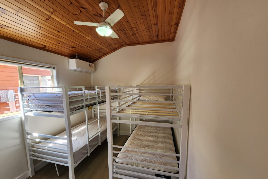 A room with two white metal bunk beds, each with mattresses. The room has a wooden ceiling with a ceiling fan and a small wall-mounted air conditioner. Sunlight filters through a window on the left wall. Walls are painted white.