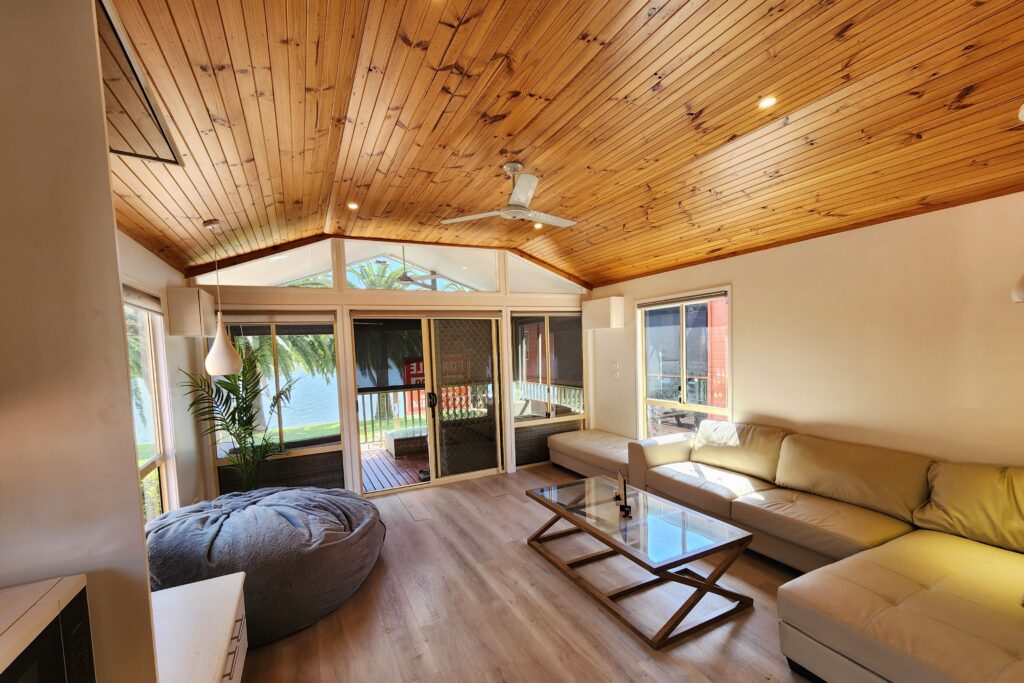 A cozy living room with a wooden ceiling and floor, featuring a large beige sectional sofa, a bean bag, and a glass coffee table. Sunlight streams in through large windows, revealing a patio with a view of trees and a body of water.
