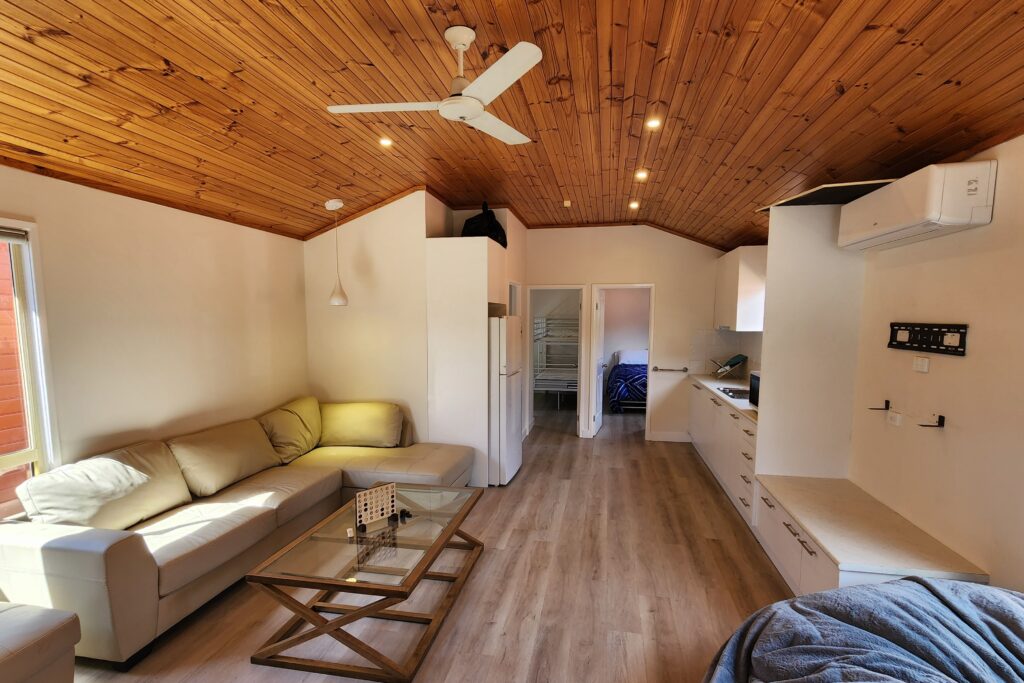 A cozy living space with wooden ceilings and light wooden floors. It features a beige sectional sofa, a glass coffee table, and a small wall-mounted TV. A ceiling fan and an air conditioning unit are visible. The space leads into a kitchen area.
