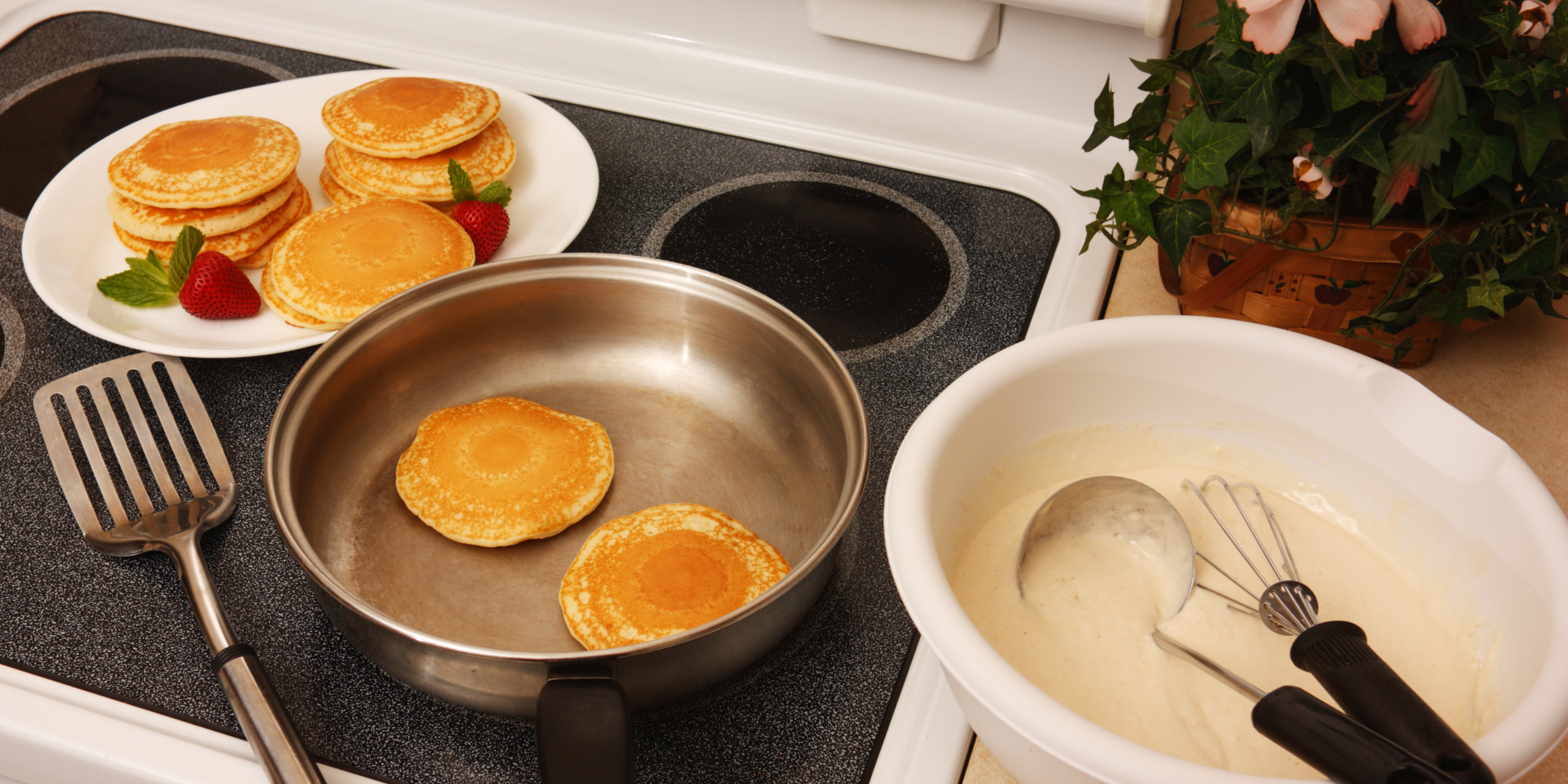 A kitchen scene showing pancakes cooking in a pan on a stovetop, with a plate of cooked pancakes and strawberries beside it. A bowl of pancake batter with a ladle and whisk are also visible. A potted plant is in the background.