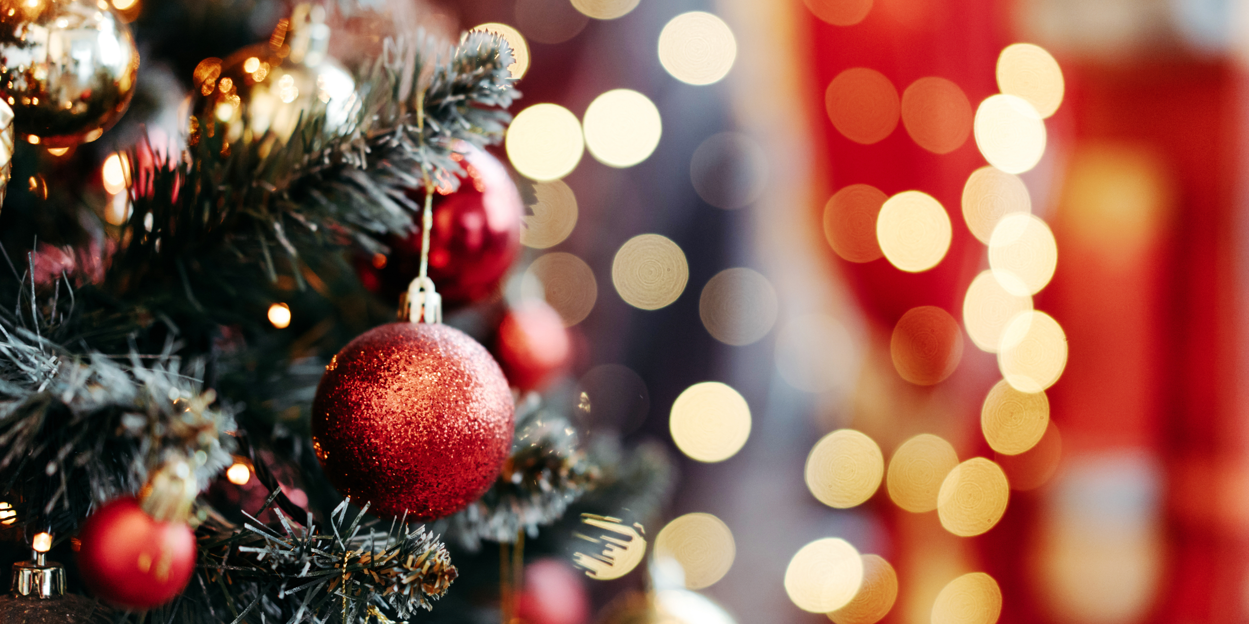 A close-up of a decorated Christmas tree with red and gold baubles. The background features soft, blurred bokeh lights in warm tones, adding a festive and cozy atmosphere to the scene.