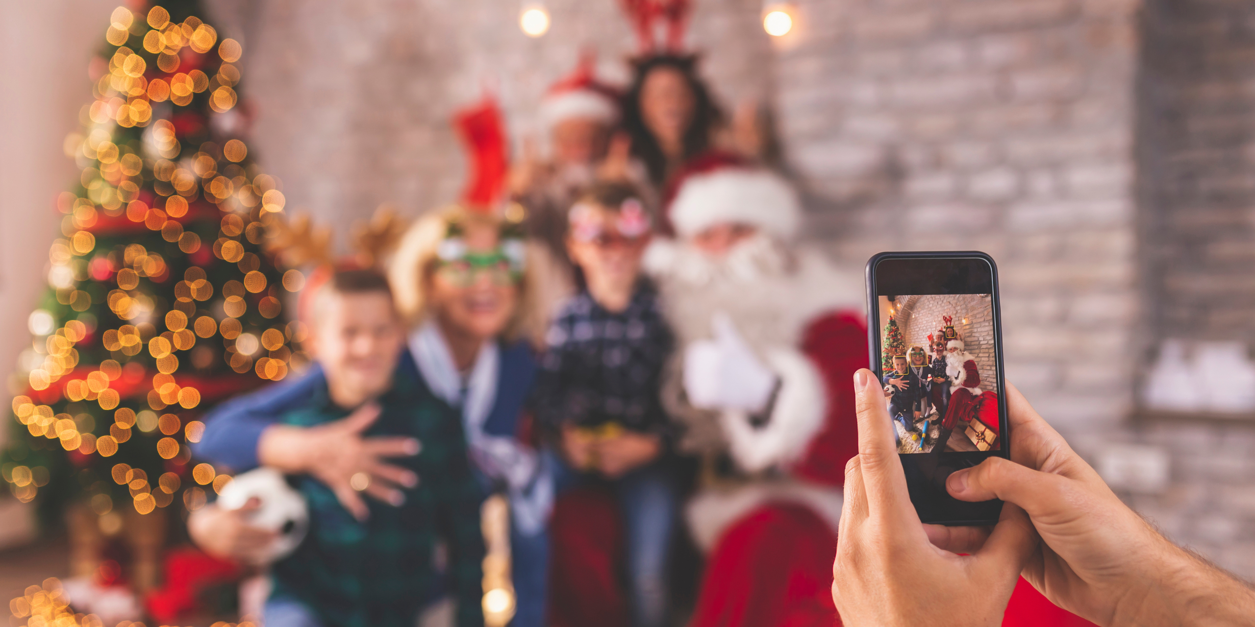 A group of people posing for a festive photo with Santa Claus. The scene is captured on a smartphone held in the foreground. They are surrounded by Christmas decorations, including a lit tree and festive outfits.