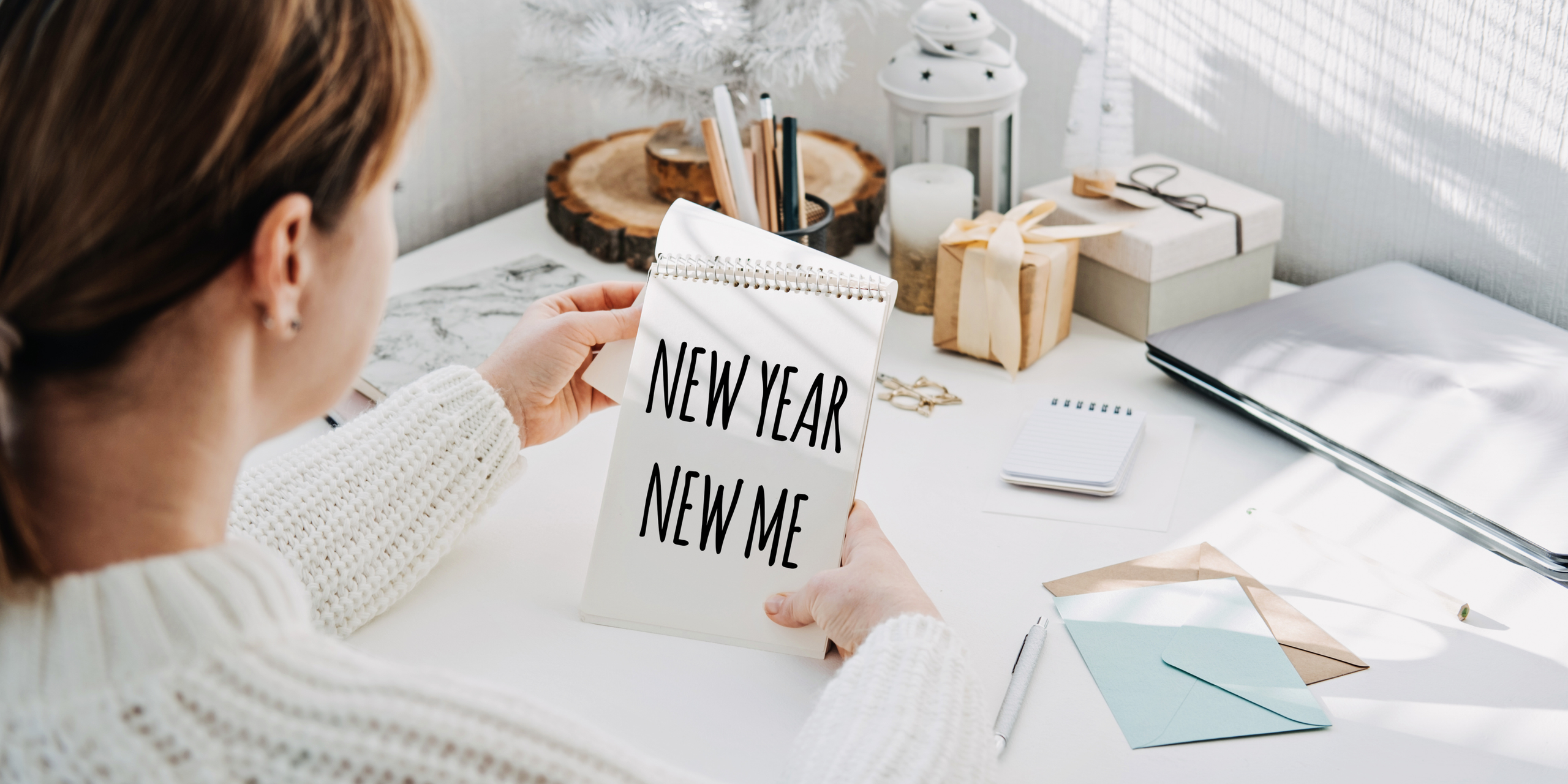 A person wearing a knitted sweater sits at a desk holding a notebook with "New Year, New Me" written on it. The desk is adorned with a small tree, a gift box, and stationery items. Sunlight filters through a window, casting shadows.