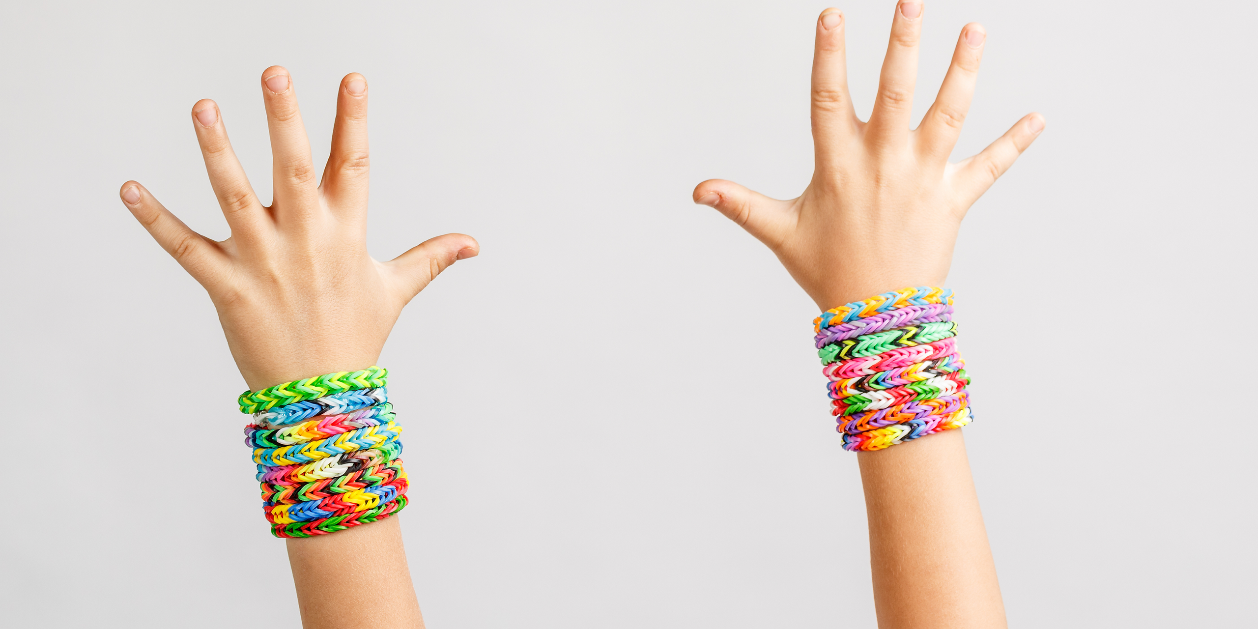 Two hands with outstretched fingers wearing colorful woven bracelets made from rubber bands. The bracelets are stacked multiple layers high, displaying a variety of bright colors against a plain white background.