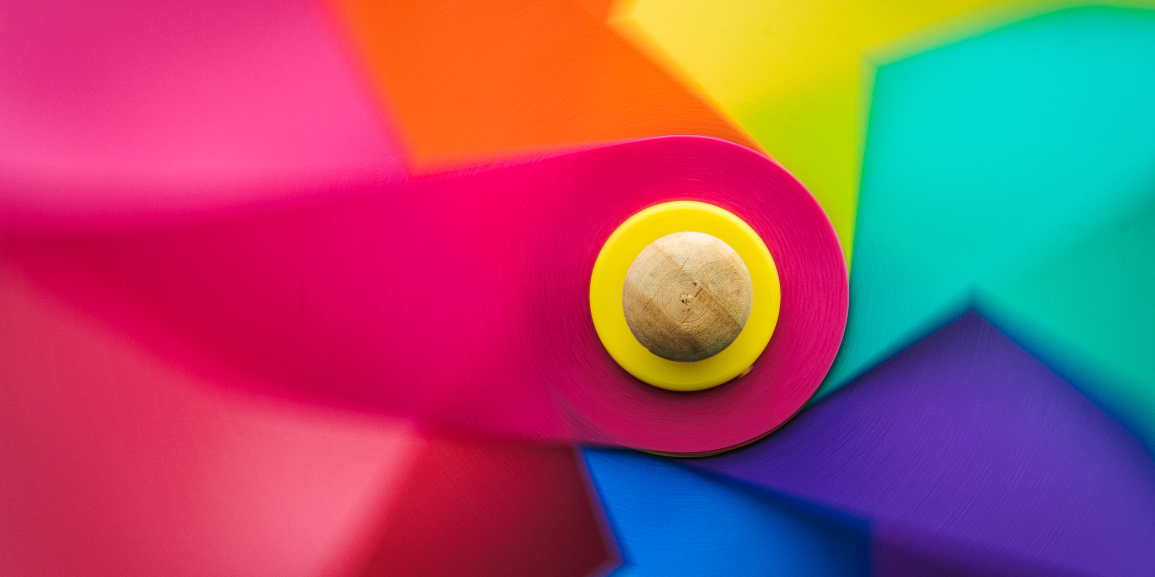 Close-up of a colorful pinwheel with vibrant hues of red, pink, orange, yellow, green, and blue radiating outward from the center. The pinwheel center is a wooden round piece framed by a bright yellow ring.