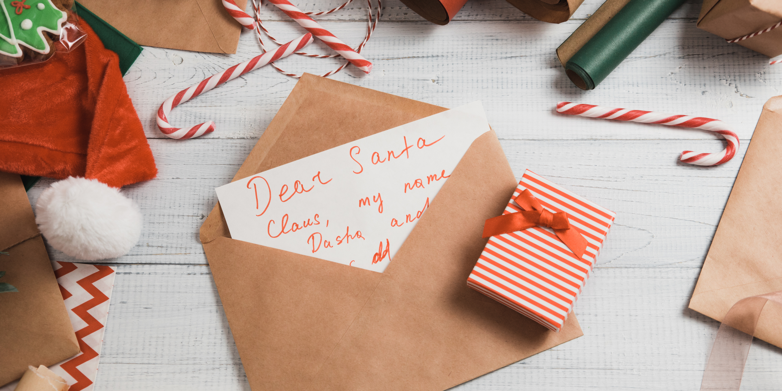 An open envelope with a "Dear Santa" letter on a white wooden table, surrounded by candy canes, Christmas cookies, and a small red and white striped gift box. Festive decorations are scattered around.