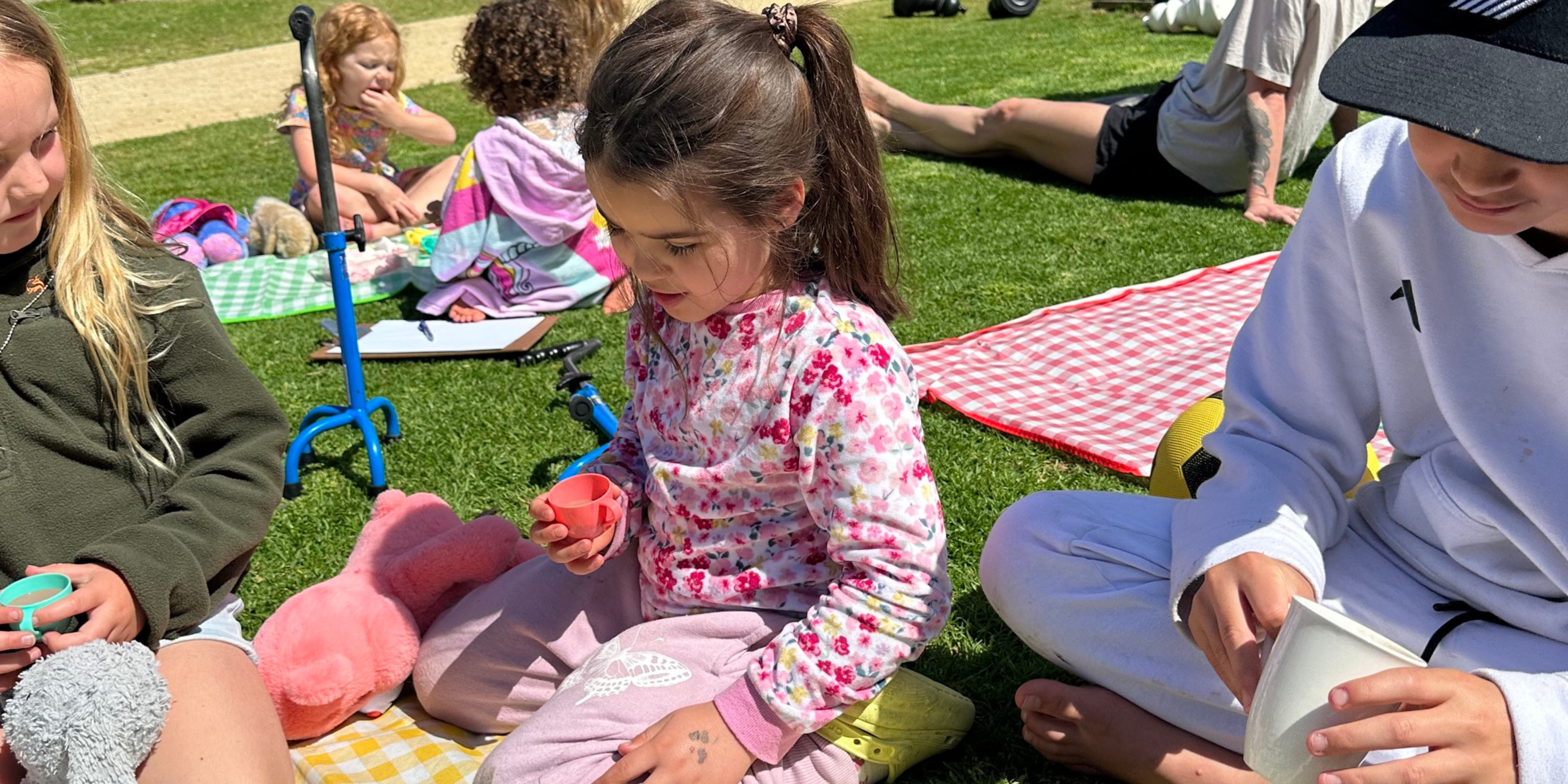 Children sitting on a grassy field with checkered picnic blankets, some holding small cups. There are scooters and toys around them. They appear to be engaged in an outdoor activity on a sunny day.