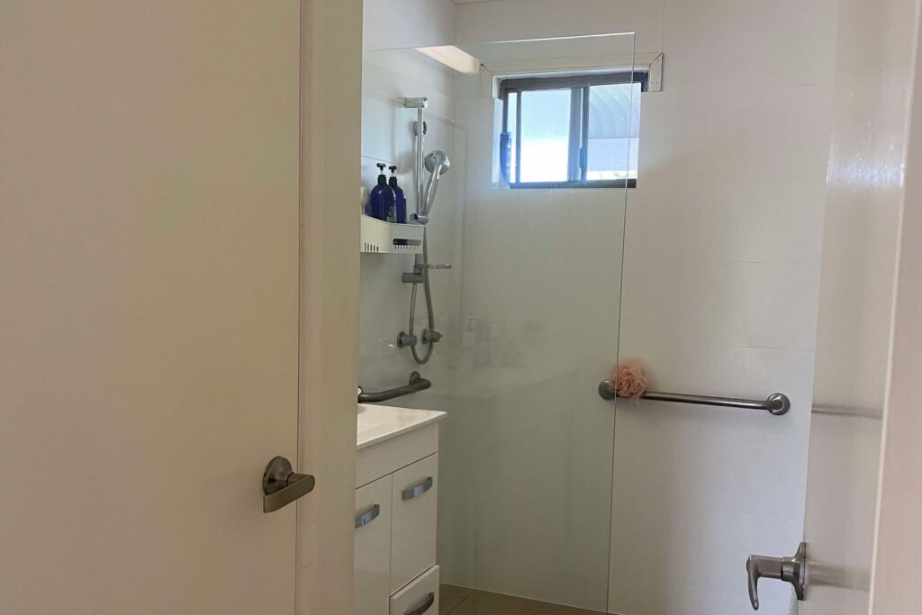 View of a bathroom featuring a glass shower with a grab bar, white vanity with a mirror, and a small window. The space has beige floor tiles and white walls, creating a minimalist and clean look.