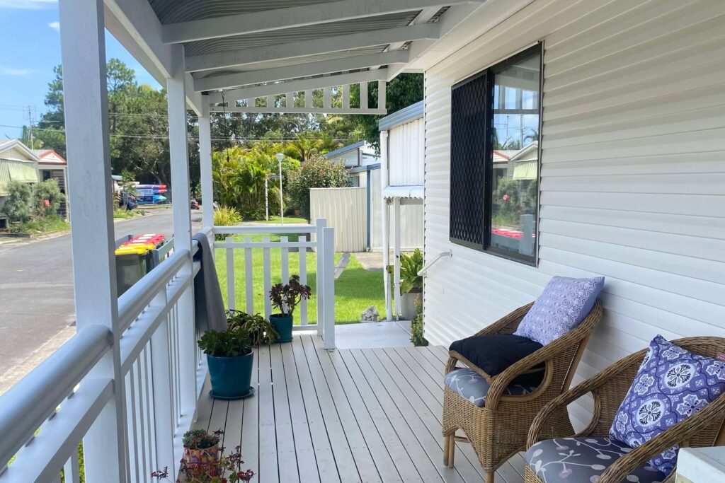 A wooden porch with wicker chairs and cushions is adorned with colorful potted plants. The space overlooks a street lined with houses and greenery. The porch has a white railing and a corrugated roof, offering a relaxing outdoor view.