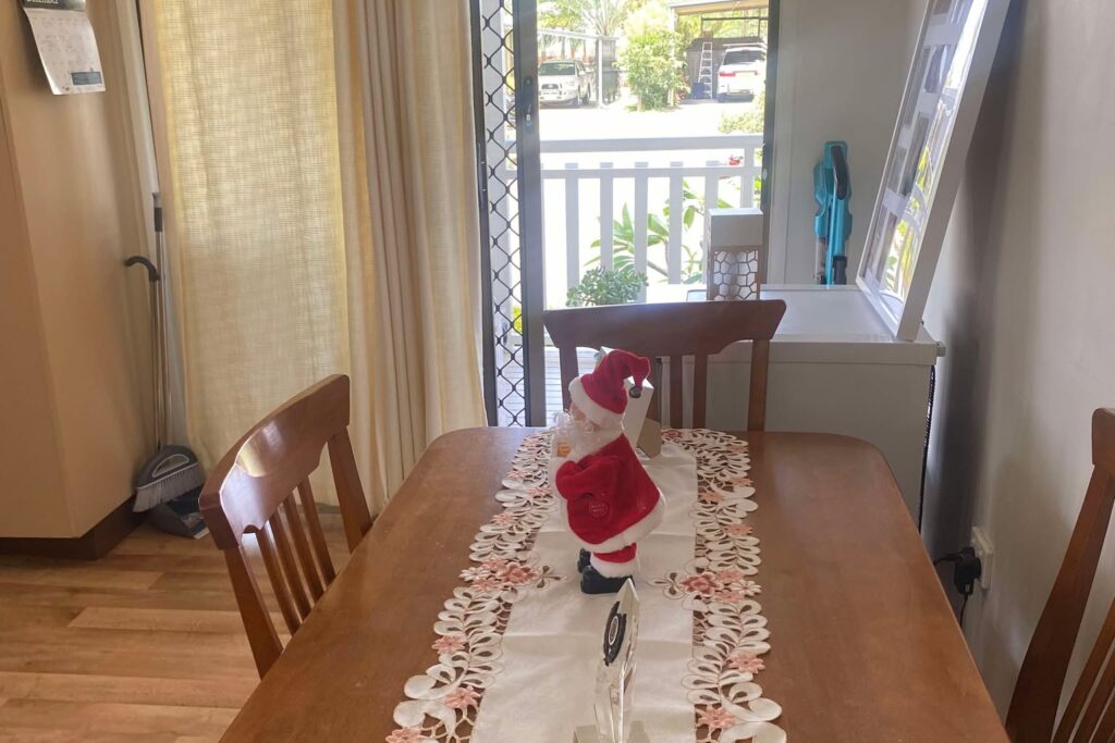 A dining room with a wooden table featuring an embroidered table runner and a Santa Claus figurine. Light brown chairs surround the table. The room has beige curtains, a window with a view of greenery, and a white side table with framed photos.