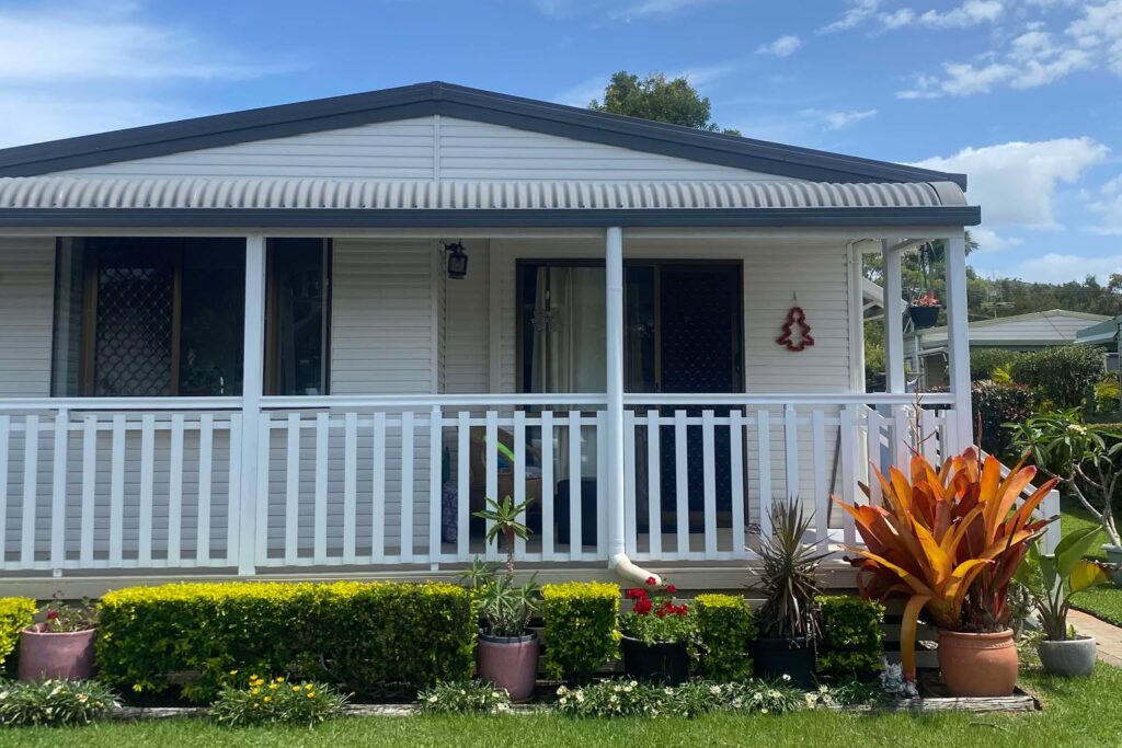 A small white house with a front porch and a blue roof. The porch is lined with white railings. Lush greenery and vibrant orange flowers adorn the front yard under a clear, blue sky.