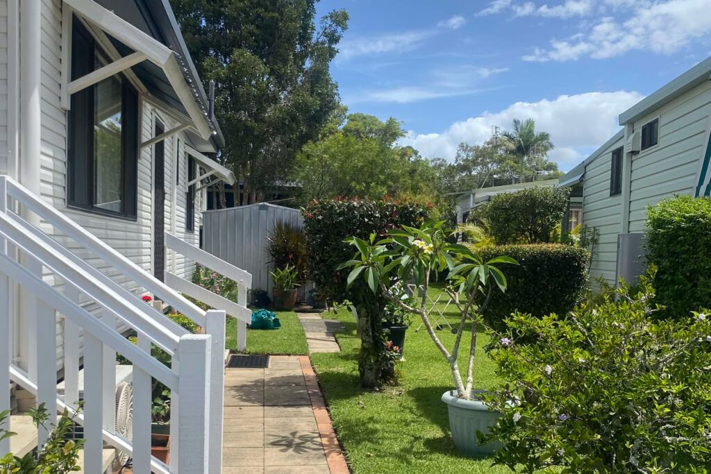 A pathway leads through a garden with lush green grass and small potted plants alongside white wooden railings. Trees and neighboring houses are visible in the background under a partly cloudy sky.