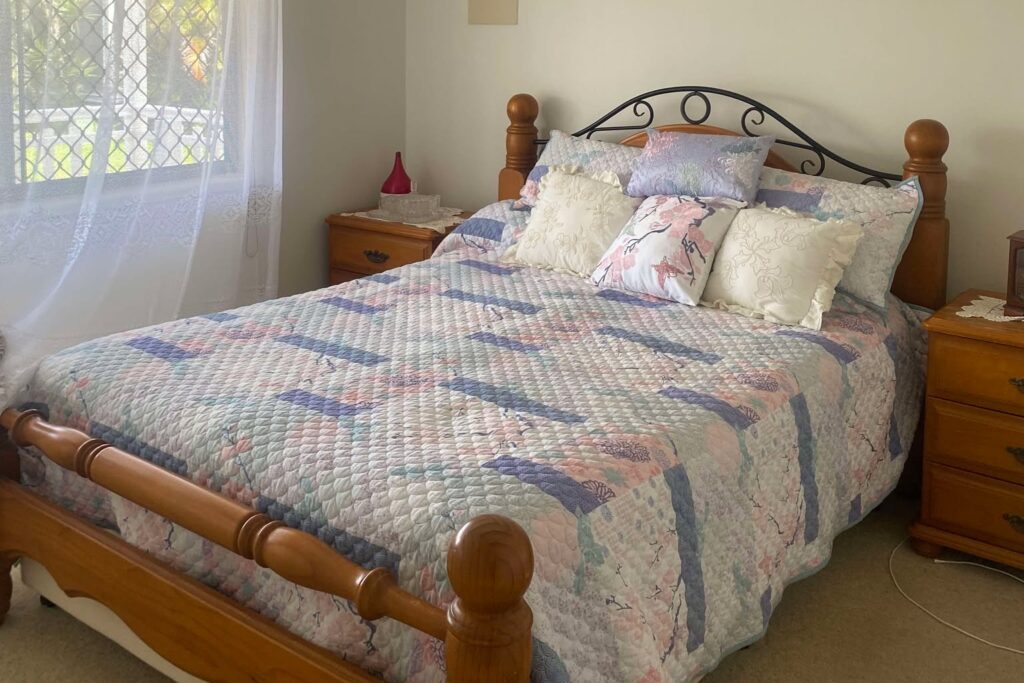 A cozy bedroom with a wooden bed frame and a quilted bedspread. The room features two wooden bedside tables, each with a lamp. A curtained window on the left lets in natural light, illuminating the light-colored walls and carpeted floor.