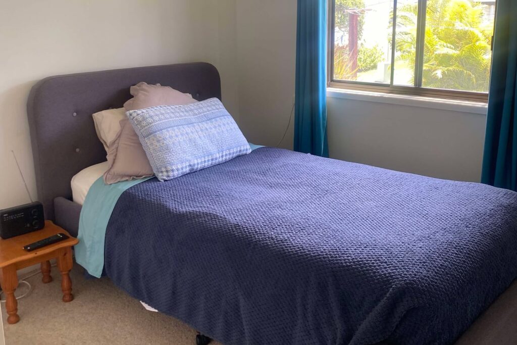 A cozy bedroom with a single bed covered in blue bedding and several pillows. A wooden nightstand with a small clock is beside the bed. The room features a window with blue curtains, offering a view of greenery outside.