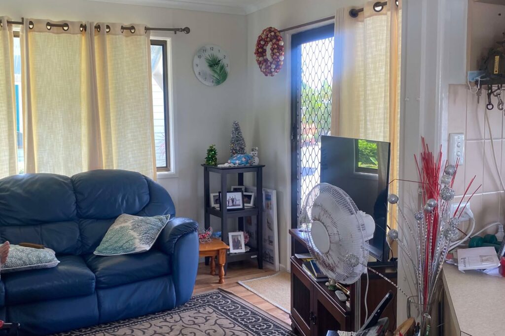 A cozy living room with a blue leather sofa, patterned rug, and curtains drawn at two windows. A TV sits on a stand with a fan on top. Various decorations are visible, including a small Christmas tree and wall clock. A sliding glass door leads outside.