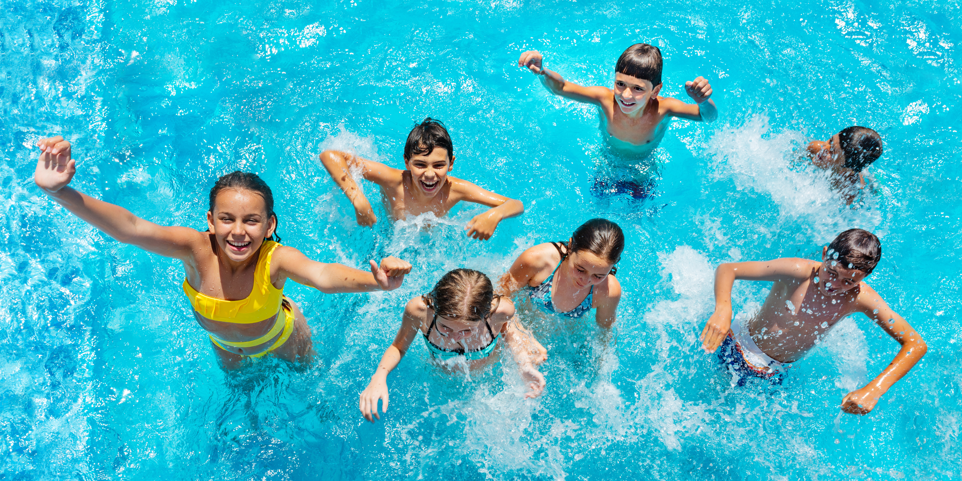 A group of seven children joyfully splashing in a clear blue swimming pool. They are smiling and appear to be having fun, with the water around them catching the sunlight.